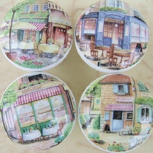 May include: Four ceramic cabinet knobs with a French cafe theme. Each knob features a different scene with a building, awning, and outdoor seating. The scenes include a restaurant, a bakery, and a cafe.