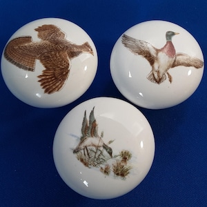 May include: Three white ceramic cabinet knobs with bird illustrations. One knob features a brown bird in flight, while the other two depict ducks. The ducks are shown in flight and near water. The knobs are round and have a glossy finish.