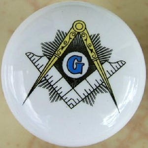 May include: A white ceramic tile with a gold and black Masonic symbol. The symbol features a compass and square with a blue letter G in the center.