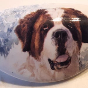 May include: A white, oblong decorative stone featuring a detailed portrait of a Saint Bernard dog. The dog has brown and white fur, with a pink tongue. Soft blue and gray watercolor-style accents are in the background.