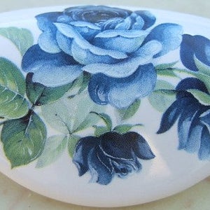 May include: A white ceramic oval dish with a blue and green floral design. The design features three blue roses and green leaves.
