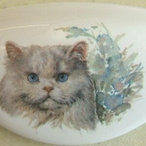May include: White ceramic decorative piece with a painted image of a fluffy gray cat with blue eyes and a floral arrangement. The cat is the focal point, set against a white background.