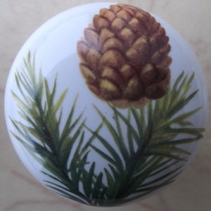 May include: White ceramic cabinet knob featuring a detailed illustration of a pine cone and green pine needles. The knob has a glossy finish and is designed for furniture or cabinet hardware. The design is in shades of brown and green.
