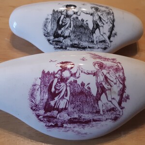 May include: Two white ceramic drawer pulls with oval shapes. Each pull features a detailed illustration of people in a garden setting. One is in black and white, the other in burgundy.