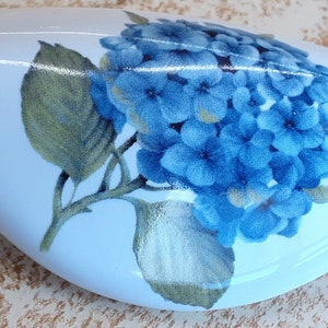 May include: A light blue ceramic vase with a floral design featuring a cluster of blue hydrangeas and green leaves.