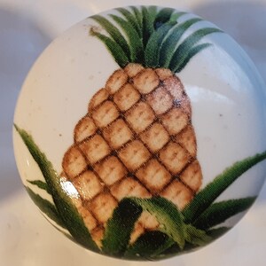 May include: A white ceramic knob with a painted image of a pineapple. The pineapple is brown and yellow with green leaves.