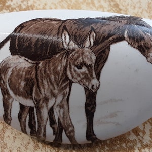 May include: A white ceramic door knob with a brown and white illustration of two donkeys. The larger donkey is standing behind the smaller donkey, and both are looking to the right.