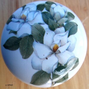 May include: White ceramic cabinet knob featuring a floral design of white magnolia blossoms and green leaves. The knob has a glossy finish and a round shape, perfect for adding a touch of nature-inspired decor to any room.
