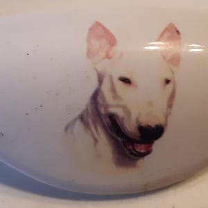 May include: White ceramic decorative piece with a painted image of a Bull Terrier dog. The dog has a white coat, pink ears, and a smiling expression. The ceramic piece is oval-shaped.