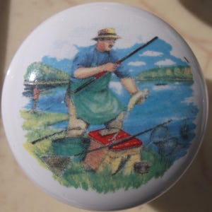 May include: White ceramic cabinet knob with a colorful illustration of a fisherman by a lake. The image shows a man in a hat and apron holding a fishing rod and a fish. The background features a blue lake and green grass.
