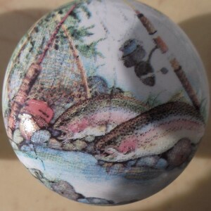 May include: A white, round ornament with a detailed illustration of a fishing scene. The image features two rainbow trout in a stream, fishing rods, and a backdrop of trees and rocks. The ornament has a glossy finish.