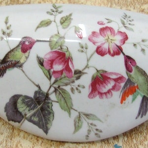 May include: A white ceramic dish with a floral design featuring hummingbirds and pink flowers. The dish has a smooth, glossy finish.