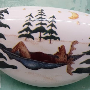 May include: A white, oval-shaped decorative stone with a painted scene of a moose relaxing in a hammock. The scene includes pine trees, a crescent moon, and stars. The moose is brown, and the hammock is blue.