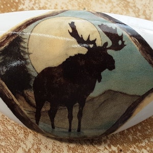 May include: White ceramic cabinet knob with a moose silhouette design. The moose is standing in front of a full moon and a forest scene.