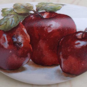 May include: Three red apples with green leaves on a white ceramic surface. The apples are arranged in a semi-circle with the largest apple in the center.