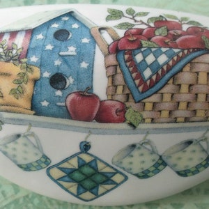 May include: A white ceramic dish with a painted scene of a birdhouse, a basket of red apples, and a potted plant. The dish has a blue, white, and red color scheme and features a whimsical, country-style design.