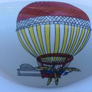 May include: White ceramic decorative piece with a colorful hot air balloon design. The balloon features a red top, yellow and white stripes, and a detailed basket. The piece has a smooth, curved shape.