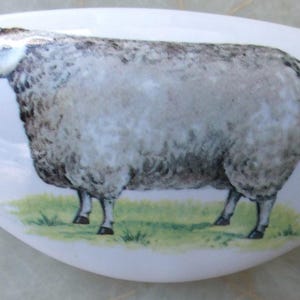 May include: A white, oblong decorative ceramic piece featuring a detailed illustration of a sheep. The sheep is depicted in shades of gray and white, standing on a patch of green grass. The piece has a glossy finish.