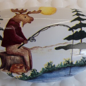 May include: White, oval-shaped decorative item with a painted scene of a moose fishing. The moose is wearing a maroon jacket and tan pants, sitting on a tree stump. A fishing rod is bent, and a yellow sun is in the background.