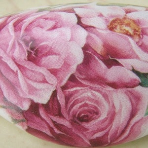 May include: A white, oval-shaped decorative object featuring a detailed floral design. The design showcases a cluster of pink roses with varying shades and textures, complemented by green leaves. The object has a glossy finish.