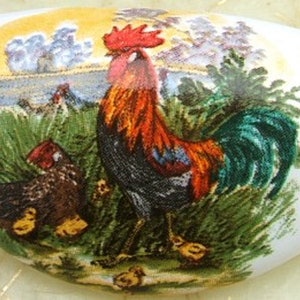 May include: A white ceramic oval-shaped dish with a rooster and hen design. The rooster is red, orange, black, and green. The hen is brown and black. The background is a yellow, green, and white landscape.