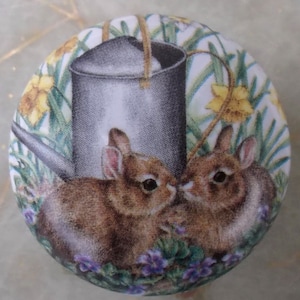 May include: A round decorative button featuring two brown bunnies nestled among purple flowers and greenery. A gray watering can is in the background, with yellow flowers. The button has a white background.
