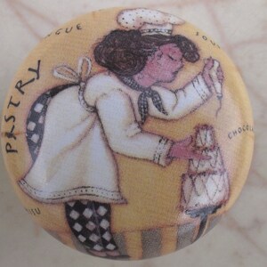 May include: A round, beige ceramic knob featuring a printed illustration of a chef in a white uniform and black-and-white checkered pants, decorating a small cake. The text "PASTRY" is printed on the left side, with other partial words visible.