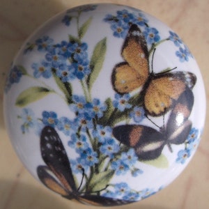 May include: A white ceramic knob with a floral design featuring blue forget-me-not flowers and two butterflies. One butterfly is orange and brown, and the other is black and brown.