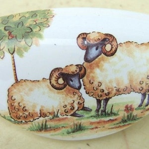 May include: A white ceramic oval dish with a hand-painted design of two sheep in a grassy field with a small tree. The sheep are brown and white with black faces and horns.