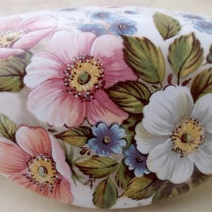 May include: White, oval-shaped decorative ceramic piece with a floral design. The flowers are pink, white, and blue, with green leaves. The piece has a glossy finish and is likely for home decor.