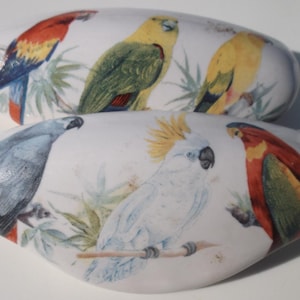 May include: Two white, oval-shaped decorative boxes with painted bird designs. The top box features a red, yellow, and blue parrot and two yellow birds. The bottom box has a blue bird, a white cockatoo, and a red and orange bird. Green foliage accents the birds.