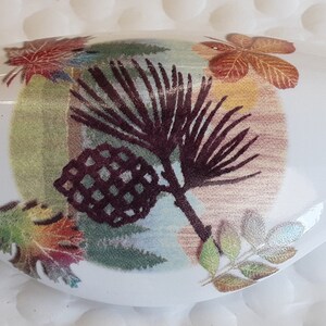 May include: White ceramic oval dish with a fall-themed design. The design features a pine branch with a pine cone, leaves in shades of red, orange, and yellow, and green foliage.