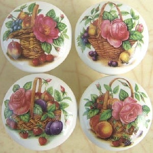 May include: Four ceramic knobs with a white background and a painted image of a basket filled with fruit and a rose. The basket is woven and brown. The fruit is red, green, and purple. The rose is pink.