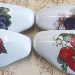 May include: Four white ceramic eyeglass cases, each decorated with a different fruit design. One case features grapes, another blackberries, a third raspberries, and the last strawberries. The fruit illustrations are in vibrant colors.