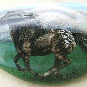 May include: A decorative, oval-shaped item featuring a running horse. The horse is depicted in shades of brown and black with white spots on its hindquarters, set against a green field and a cloudy sky. The item has a glossy finish.