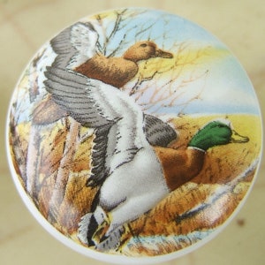 May include: A round, white ceramic knob featuring a detailed illustration of three ducks in flight. The ducks have brown, white, and green plumage, set against a backdrop of a field and a blue sky with bare trees. The knob is likely for a cabinet or drawer.