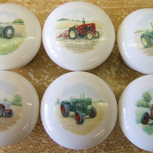 May include: Six white ceramic knobs with different tractor designs. Each knob features a different colored tractor with a green, red, or gray body. The tractors are depicted in a rural setting with green fields and blue skies.