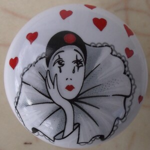 May include: A white ceramic knob with a black and white illustration of a clown with a red nose and red hearts around the edge.