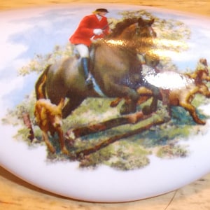 May include: White, oval-shaped porcelain decorative piece with a hunting scene. The image depicts a rider in a red coat on a brown horse jumping over a fence, with dogs running alongside. The background shows trees and a blue sky.