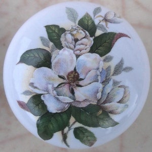 May include: A white ceramic cabinet knob featuring a detailed floral design. The design includes large white magnolia blossoms with green leaves and a brown center. The knob is round and has a glossy finish.