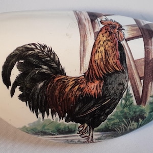 May include: A white, oval-shaped ceramic decorative piece featuring a colorful rooster illustration. The rooster has black, brown, and orange feathers, standing in front of a wooden fence and green grass.