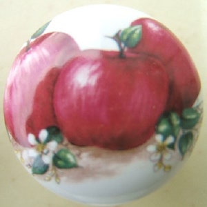 May include: A white ceramic knob with a painted design of three red apples and green leaves. The apples are arranged in a cluster with the largest apple in the center.