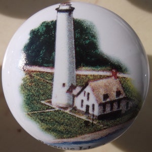 May include: A white ceramic knob with a painted image of a lighthouse and a house. The lighthouse is tall and white with a black roof. The house is white with a brown roof and windows. The background is green grass and blue sky.
