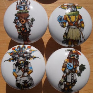 May include: Four white ceramic cabinet knobs with colorful illustrations of Native American Kachina figures. Each knob features a different design, showcasing detailed artwork and vibrant colors. These decorative knobs add a unique touch to furniture.