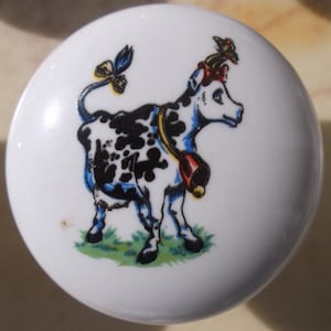 May include: White ceramic cabinet knob featuring a cartoon cow with black spots, a red bell, and a blue bow. The cow is standing on green grass. The knob is round and has a glossy finish.