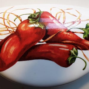 May include: A white, oval-shaped decorative object featuring a vibrant illustration of several red chili peppers with green stems. The peppers are arranged with dried, golden-brown strands.