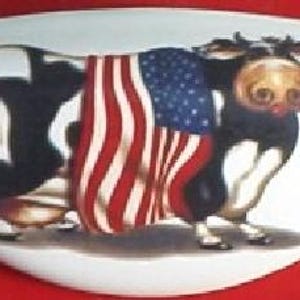 May include: A white, oval-shaped decorative plate featuring a cartoon cow draped in an American flag. The cow is black and white with a large, expressive face. The flag covers the cow's back, with red and white stripes and a blue field of stars.
