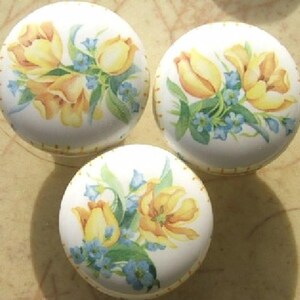 May include: Three white ceramic cabinet knobs with a floral design. Each knob features yellow tulips and blue forget-me-nots. The knobs have a yellow dotted border and are set against a neutral background.
