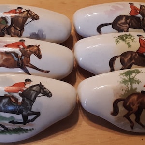 May include: Six white ceramic drawer pulls with a horse and rider design. Each pull features a different horse and rider in a variety of colors, including brown, black, and white.