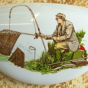 May include: White ceramic decorative piece with a fishing scene. The illustration depicts a person fishing with a fishing rod, net, and tackle box. The person is wearing a hat, jacket, and boots. The background includes water and grass.